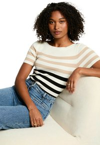 Haut en jersey à manches trois quarts dans des rayures crème, beige et noir, associé à un jean taille haute bleu. Le modèle pose sur un canapé de couleur claire.