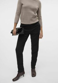 Pantalon noir sur mesure avec une texture lisse, associé à un pull ajusté beige clair. Tient un sac à main noir et porte des bottines à talons marron foncé.