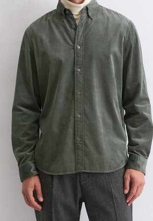 Mann trägt ein olivgrünes, zugeknöpftes Cordhemd über einem weißen Rollkragenpullover und grauen Wollhosen.