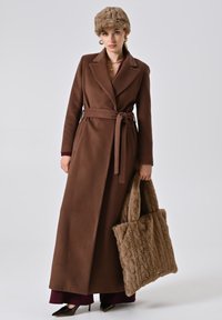 Bruine lange jas met riem en zakken, gecombineerd met een getextureerde tan handtas. Model draagt een bijpassende bruine hoed en hoge hakken.