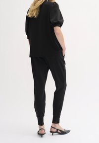 Schwarzes, lässig sitzendes Oberteil mit Puffärmeln, kombiniert mit passender, schmal zulaufender Hose. Die Schuhe bestehen aus schwarzen Riemchen-Absätzen mit spitz zulaufender Zehenpartie.