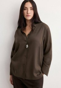 Vrouw met lang donker haar, gekleed in een losse bruine blouse met knopen en een hangertje aan een ketting, staand met één hand in haar zak tegen een witte muur.