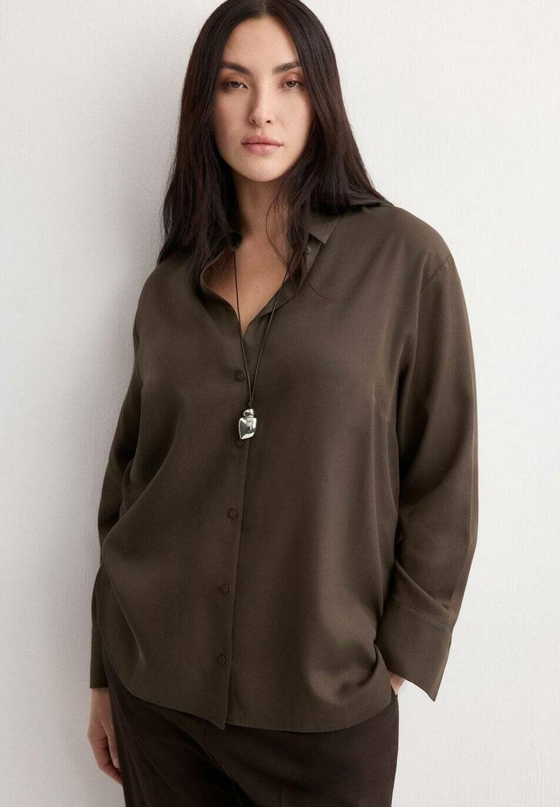 Vrouw met lang donker haar, gekleed in een losse bruine blouse met knopen en een hangertje aan een ketting, staand met één hand in haar zak tegen een witte muur.