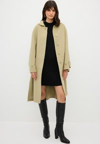 Cappotto in misto lana beige con cappuccio, chiusura a bottoni, tasche laterali e maniche oversize, indossato sopra un vestito nero a coste e stivali neri sopra il ginocchio.