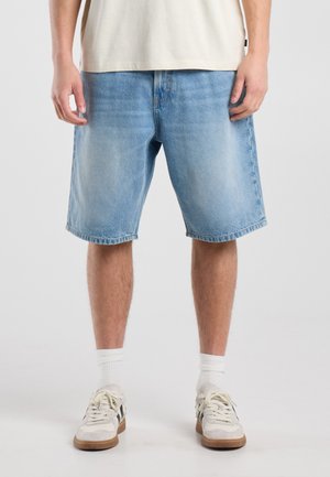 Persoon draagt lichtblauwe denim shorts, witte sportsokken, witte sneakers en een beige shirt dat net boven de taille is geknipt.