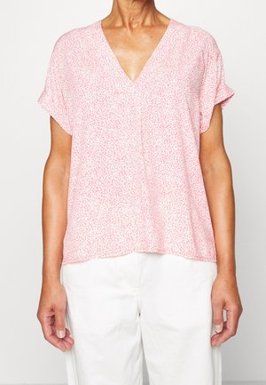 Vrouw draagt een lichtroze blouse met korte mouwen en een V-hals met witte vlekjes, gecombineerd met een witte broek, tegen een effen achtergrond.