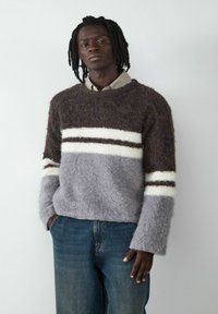 Flauschiger, mehrfarbiger Pullover mit braunen, grauen und weißen Streifen. Runder Ausschnitt, lockere Passform und strukturierter Stoff. Kombiniert mit blauen Jeans.