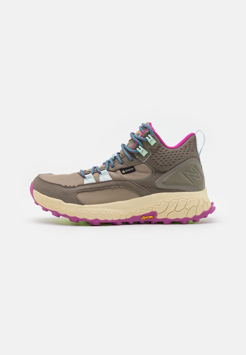 Zapato de senderismo con parte superior de textil marrón, acentos en rosa y cordones azules. Presenta una entresuela gruesa y texturizada en beige y una suela de goma vibrante.