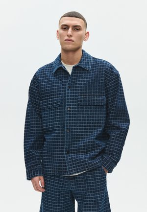 Mladić s kratkom kosom, nosi mornarsko plavu jaknu na kockice i odgovarajuće hlače, stoji nasuprot jednobojnoj svijetloj pozadini.