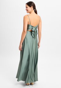 Langes, fließendes grünes Satin-Kleid mit einem gebundenen Rückendesign, dünnen Trägern und einer glatten Textur. Es hat ein tailliertes Oberteil und einen voluminösen Rock.