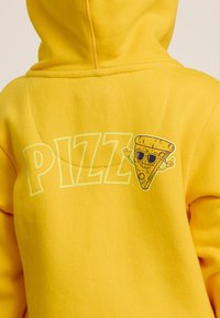 Un sweat à capuche jaune avec un graphique d'une part de pizza souriante portant des lunettes de soleil et le mot "PIZZ" en lettres grasses. Texture douce.