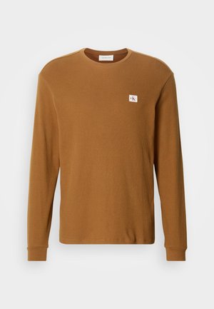 Braunes langärmliges Baumwollhemd mit gerippter Textur, Rundhalsausschnitt und kleinem weißen Logo-Patch auf der oberen linken Seite.