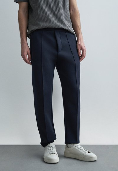 Navy tailored pants with side stripes, soft fabric, and a relaxed fit, paired with white sneakers. Minimalist design and clean lines.