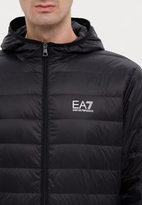 Giacca imbottita nera con cappuccio e zip frontale. Il materiale ha una superficie liscia con sezioni trapuntate e un logo bianco sul petto.
