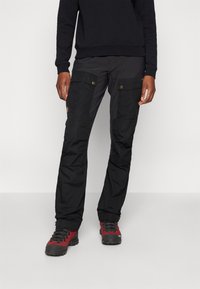 Black outdoor pants with a cargo design, featuring multiple pockets, button closures, and a durable texture, paired with red hiking shoes.