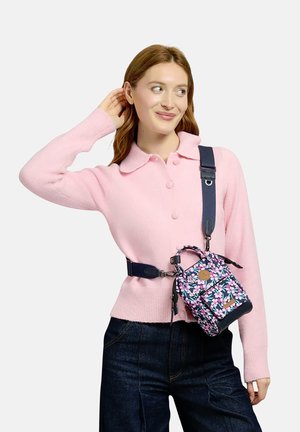 Frau trägt eine rosa Strickjacke und dunkle Jeans und hat eine kleine florale Umhängetasche mit schwarzem Riemen über Schulter und Taille.