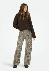 Junge Frau, die steht und einen dunkelbraunen Pullover sowie weite, beige Hosen mit Leopardenmuster und schwarzen Schuhen trägt, während sie zur Seite schaut.