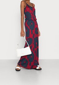 Robe maxi rouge et bleu marine avec un motif floral, fabriquée en tissu doux. Associée à un sac à main blanc et des sandales noires avec des bretelles blanches.