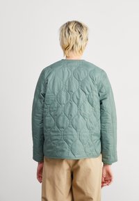 Gequilted jas van zacht groene stof, met een ronde halslijn en een gewatteerd golfpatroon op de achterkant. Gecombineerd met beige broek.