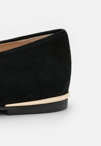 Chaussure plate en daim noir avec un talon et un fin accent en métal doré au-dessus d'une semelle noire basse sur fond blanc.