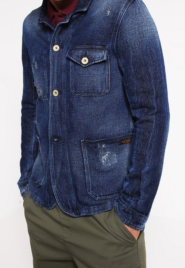 Denim jacket in dark blue with subtle fading, two chest pockets, and a slightly distressed texture. Features button closures and a collared design.