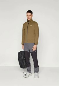 Giacca olive green con zip, dalla texture liscia, abbinata a pantaloni grigio-neri. Il modello tiene uno zaino nero e indossa sneakers grigie chiare.