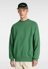 Sudadera verde de cuello redondo en suave tejido de algodón, con hombros caídos y puños acanalados. Combinada con una gorra negra que muestra un logotipo verde.