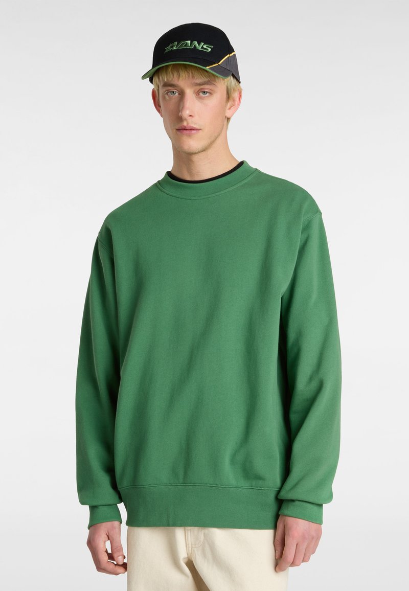 Sudadera verde de cuello redondo en suave tejido de algodón, con hombros caídos y puños acanalados. Combinada con una gorra negra que muestra un logotipo verde.