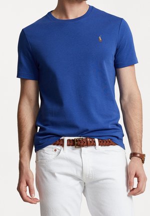 Mann trägt ein blaues kurzärmeliges T-Shirt mit kleinem Logo, weiße Hose, einen braunen geflochtenen Gürtel und eine braune Armbanduhr.