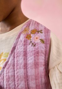 Lila rutiga västen med blommönsterbroderi på axeln, bärs över en ljusfärgad skjorta med strukturerad tyg. Mjuk, lager-på-lager design.