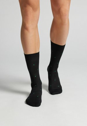 Beine in schwarzen Socken mit kleinen grünen Rautenmuster, stehend auf einem hellgrauen, glatten Boden.