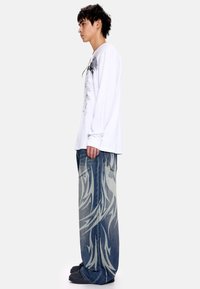 Άσπρο πουκάμισο με μακριά μανίκια και γραφικό τύπωμα, σε συνδυασμό με ανοιχτό μπλε φαρδιά τζιν με αφηρημένα γκρι σχέδια. Μαύρα παπούτσια χωρίς κορδόνια.