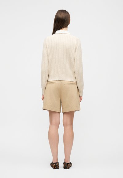 Femme portant un pull crème côtelé à manches longues, un short beige jusqu'aux genoux, et des ballerines à imprimé léopard, vue de dos sur un fond uni.