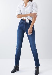 Mujer con jeans ajustados azul oscuro, camisa blanca de botones anudada con las mangas remangadas y botines negros de tacón, posando contra un fondo blanco.