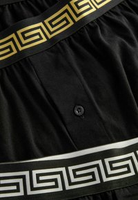 Schwarzer Stoff mit plissierter Textur, ausgestattet mit einem gold-schwarzen geometrischen Muster am Taillenbund. Enthält ein schwarzes Knopfdetail.