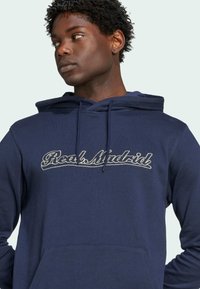 Marinblå huvtröja i en bomullsblandning, med en framficka i känguru-stil och kontrasterande broderad text över bröstet.