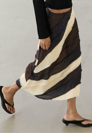 Persona che cammina con una maglia nera a maniche lunghe, gonna lunga a righe nera, bianca e marrone e sandali neri con tacco su un pavimento liscio.