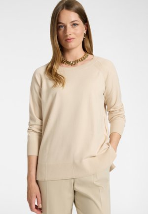 Donna con lunghi capelli castano chiaro indossa maglione e pantaloni beige, collana con catena d'oro e orecchini a cerchio dorati, posando con una mano in tasca.