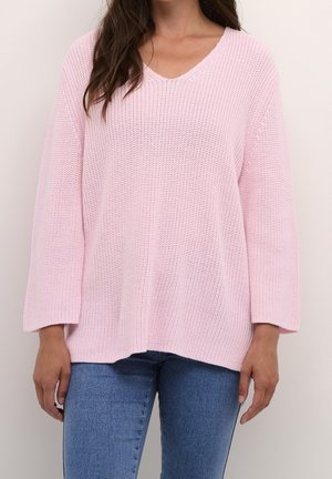 Femme portant un pull tricoté ample de couleur rose clair à manches longues et un jean bleu, debout devant un fond uni clair.
