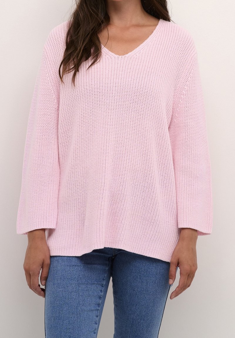 Femme portant un pull tricoté ample de couleur rose clair à manches longues et un jean bleu, debout devant un fond uni clair.