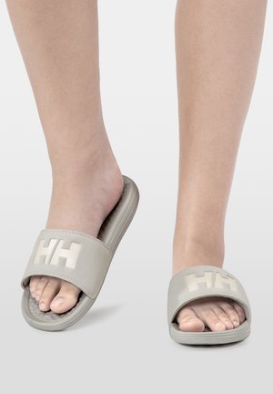 Stopy w beżowych wsuwanych sandałach z logo "HH" na paskach, jedna stopa lekko uniesiona, ukazująca teksturę podeszwy sandała.