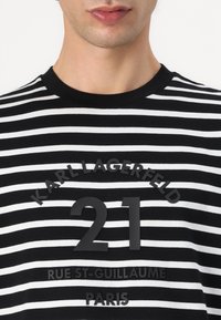 Juostuota juoda ir balta marškinėliai su apvaliu iškirpimu, guminis logotipas ir juodas tekstas, nurodantis "Karl Lagerfeld 21 Rue St-Guillaume Paryžius."