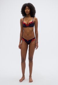 Σετ εσωρούχων σε ναυτικό μπλε με κόκκινα δαντελένια στοιχεία. Το σουτιέν διαθέτει μπανέλα και ρυθμιζόμενους ιμάντες· τα ταιριαστά σλιπ σε στυλ μπικίνι έχουν παρόμοια δαντελένια διακόσμηση.