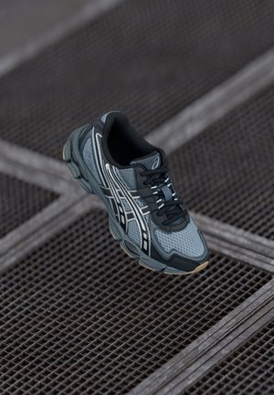 Chaussure de sport grise et noire avec des détails blancs flottant au-dessus d'une surface en grille métallique.