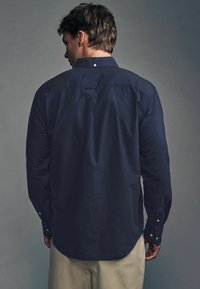 Tengerészkék, hosszú ujjú ing, gombos gallérral, egyenes szabású, lekerekített szegéllyel. Pamut anyag, sima tapintással, gombos mandzsettával.