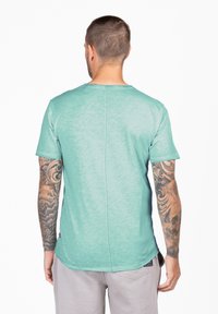 T-shirt a maniche corte di un leggero color teal con una texture sottile, scollo rotondo e cuciture a contrasto, che mette in mostra i tatuaggi sulle braccia.