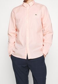 Camicia button-up di cotone rosa chiaro con colletto a punte, maniche lunghe e un piccolo logo verde in alto a sinistra.