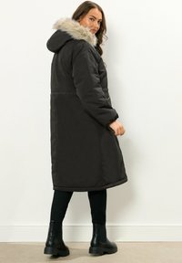 Pitkä musta untuvatakki, jossa on turkiskauluri, vyötärö on kireä ja sivutaskut, käytetty mustien leggingsien ja nilkkasaappaiden kanssa.