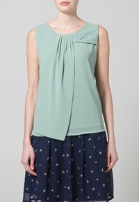 Top senza maniche color verde menta con design plissettato e dettaglio a fiocco. Abbinato a una gonna blu navy con piccoli motivi di uccelli bianchi. Tessuto dalla consistenza morbida.