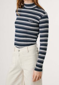 Marinblå och vita randiga långärmade överdelar med hög hals. Ribbad textur och liten logotyp på det övre vänstra. Kombinerat med ljusa denim.